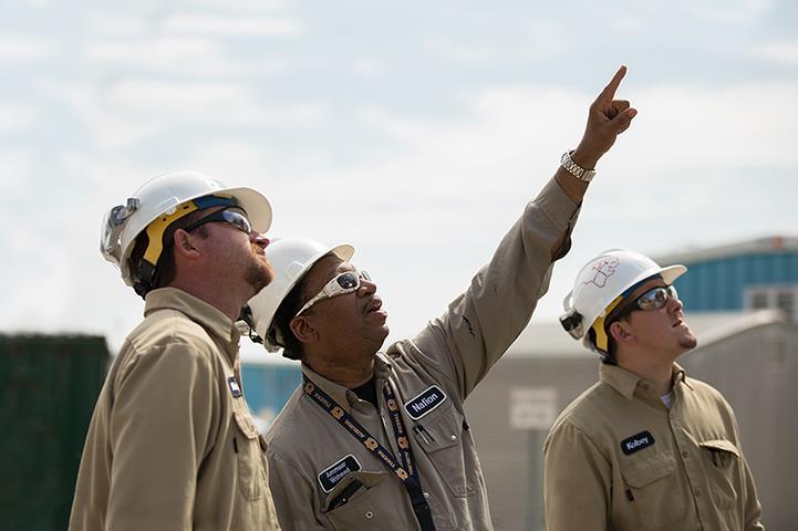 three men in white hardhats tan coveralls and sun glasses looking up at something the middle man is