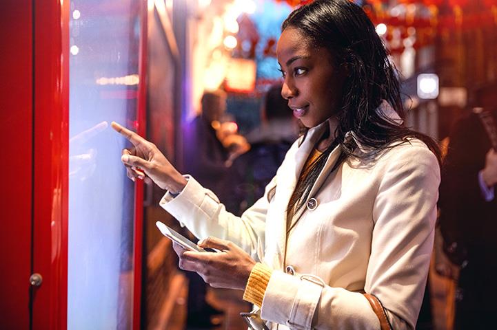 An image of a woman standing in front of a touchscreen holding a phone.