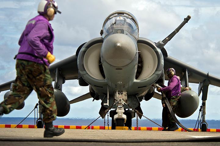 one man in bdu walking in front of a harrier jet while another man adds fuel
