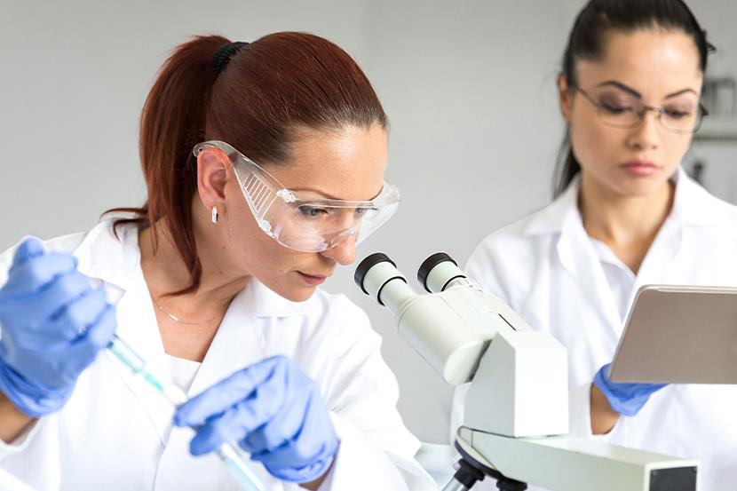 woman looking into a microscope while holding a syringe another woman looking at a tablet